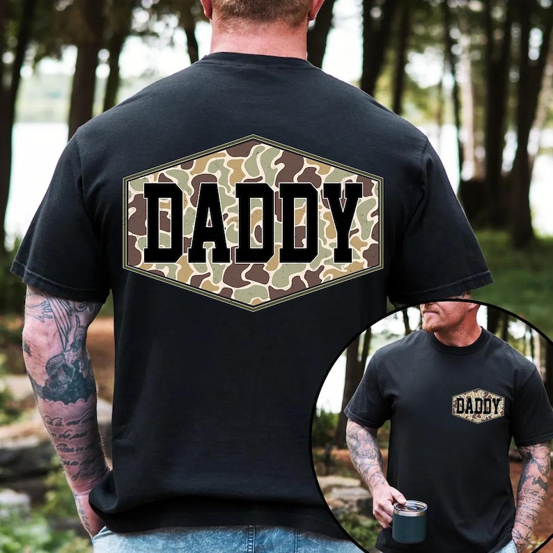 May include: Black t-shirt with a camouflage design in a hexagonal shape on the back, featuring the word "DADDY" in bold, black letters. The shirt is short-sleeved and appears to be made of cotton.