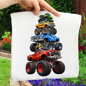 May include: A white cushion cover displaying a stack of five vibrant monster trucks. The trucks are in red, blue, yellow, and green, each with large tyres and detailed designs. The cushion cover is held up by a hand.