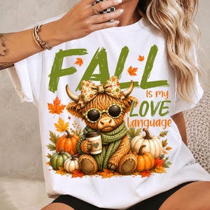 May include: A white t-shirt featuring a graphic of a highland cow wearing a floral bow, sunglasses, and a green scarf while holding a coffee cup. The text reads FALL is my LOVE Language, surrounded by orange and green autumn leaves and pumpkins.