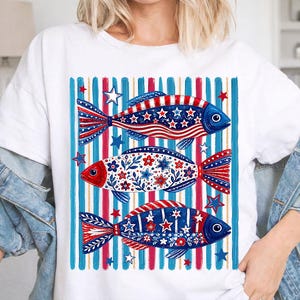 May include: White t-shirt featuring a patriotic design of three fish with red, white, and blue patterns. The fish are set against a backdrop of blue and red stripes and stars, creating a festive and eye-catching design.