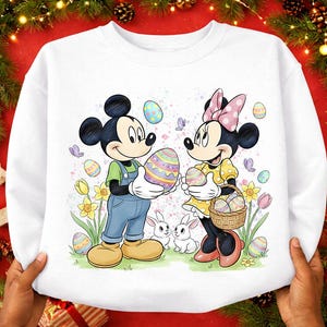 May include: White sweatshirt featuring Mickey and Minnie Mouse holding Easter eggs. The design includes colorful eggs, butterflies, flowers, and rabbits. Mickey wears overalls, and Minnie wears a yellow dress. The overall theme is Easter.