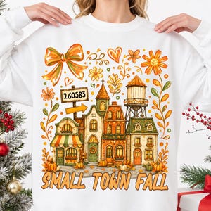 Op de afbeelding: Een wit sweatshirt met lange mouwen en een kleurrijke afbeelding van een klein stadje in herfstthema, met gebouwen, een watertoren, pompoenen en bloemen. De tekst "260583" staat op een wegwijzer en "SMALL TOWN FALL" staat eronder gedrukt.