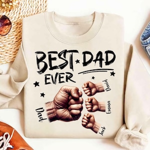 May include: Cream-colored sweatshirt with the text "BEST DAD EVER" in black, accompanied by illustrations of fists and stars. The names "Dad," "David," "Emma," and "Jack" are printed vertically near the fists.