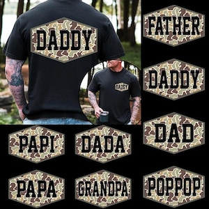 May include: Black t-shirts with the word "DADDY" in bold, black letters inside a camouflage hexagon. Other shirts display "FATHER", "DAD", "PAPI", "DADA", "GRANDPA", and "POPPOP" in a similar style.