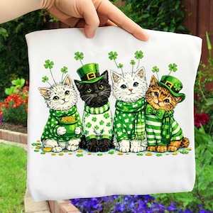 May include: White fabric featuring four cartoon cats dressed for St. Patrick's Day. Each cat wears a green hat with a shamrock, green clothing, and a gold coin. The cats are in various colors: white, black, and orange.