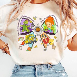 May include: Cream-colored t-shirt featuring a colorful bow design with a daisy center. The bow includes illustrations of a dragon, a woman, a school bus, and other elements. Text on the bow reads "Take chances make mistakes get messy."