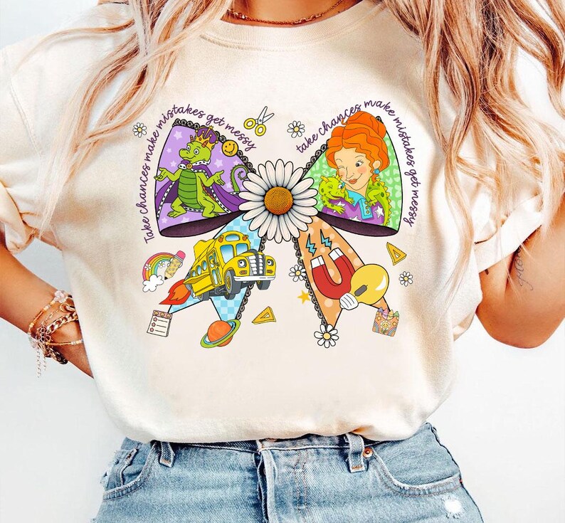 May include: Cream-colored t-shirt featuring a colorful bow design with a daisy center. The bow includes illustrations of a dragon, a woman, a school bus, and other elements. Text on the bow reads "Take chances make mistakes get messy."