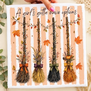May include: A white fabric square featuring a peach and white striped background with the text A girl's gotta have options. Four decorative brooms are displayed, each adorned with autumn leaves, berries, and bows in orange, purple, and green.