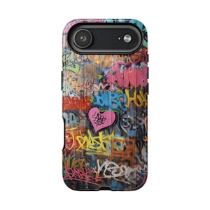 May include: A phone case featuring a vibrant graffiti design. The case is covered in colorful spray-painted tags, with a prominent pink heart in the center. The design includes various colors like blue, yellow, and pink, creating a street art aesthetic.