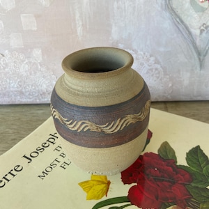 May include: A small, handcrafted ceramic vase with a beige base and a dark brown band featuring a wavy, lighter brown design. The vase has a wide opening and a slightly flared rim. The vase is sitting on a book with floral illustrations.