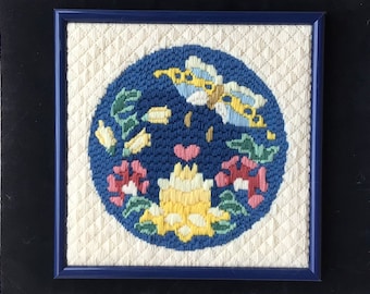 May include: A framed needlepoint embroidery featuring a blue circle with a yellow butterfly, pink and yellow flowers, and green leaves on a white background.