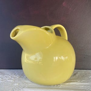 May include: A pale yellow ceramic pitcher with a rounded body and a curved handle. The pitcher has a spout for pouring and a glossy finish. The background is a dark purple color. The pitcher is sitting on a white lace tablecloth.