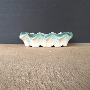 May include: A rectangular ceramic dish with a wavy, geometric design. The dish is primarily white with accents of light blue and teal. The edges have a scalloped appearance. The dish is sitting on a neutral-toned surface.