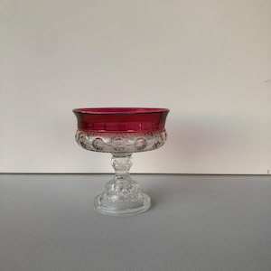May include: A clear glass pedestal bowl with a ruby red rim. The bowl has a decorative, patterned design on the base and around the rim. The stem is also clear glass with a detailed, textured pattern. The bowl is likely used for serving desserts or displaying small items.