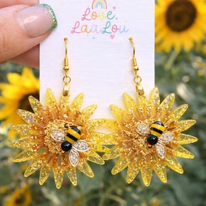 May include: A pair of yellow sunflower dangle earrings with gold-colored hooks. Each earring features a yellow flower with glitter and a bee charm with black and yellow stripes. The text "Bee My Sunflower Dangles" is at the bottom.