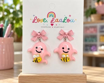 May include: A pair of pink ghost-shaped earrings on a white display card labeled Love, LaaLou. Each ghost features a small yellow and black bee on its front and is topped with a pink bow. The design includes black eyes and a smile.