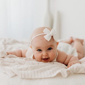 Può includere: Un bambino sorridente con una fascia bianca con fiocco, sdraiato su una coperta di maglia color crema. Il bambino indossa un vestito bianco e guarda direttamente la fotocamera.