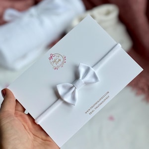 Könnte beinhalten: Ein weißes Baby-Haarband mit einer weißen Schleife. Das Haarband befindet sich auf einer weißen Karte mit dem Text "That's So Lovely" und der Website-Adresse "www.thatssolovely.com".