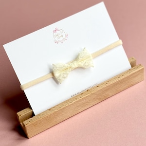 Pode incluir: Uma faixa de cabelo com laço de renda creme exibida em um cartão branco dentro de um suporte de madeira. O cartão tem um logotipo que diz "That's So Lovely" e o endereço do site. O fundo é rosa claro.