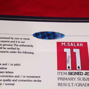 May include: A white card with black text stating that the item has been examined by experts and is authentic. The card also includes a photo of a red and white soccer jersey with the number 11 and the name "M. Salah" printed on it. The text "ITEM: SIGNED JERSEY", "PRIMARY SUBJECT: M. Salah", and "RESULT/GRADE: AUTHENTIC" is printed on the card.