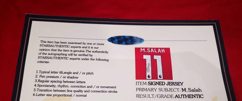 May include: A white card with black text stating that the item has been examined by experts and is authentic. The card also includes a photo of a red and white soccer jersey with the number 11 and the name "M. Salah" printed on it. The text "ITEM: SIGNED JERSEY", "PRIMARY SUBJECT: M. Salah", and "RESULT/GRADE: AUTHENTIC" is printed on the card.