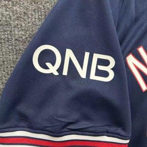 May include: Close-up of a navy blue jersey sleeve with the white letters "QNB" prominently displayed. The sleeve has a white, red, and white striped trim at the bottom. The jersey appears to be made of a breathable fabric.