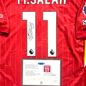May include: A red soccer jersey with the name "M. SALAH" and the number "11" on the back. The jersey has a white signature on the number 1 and a white logo on the number 1. The jersey has a white tag on the bottom with the text "THE REAL DEAL" and "AUTHENTIC" in black.