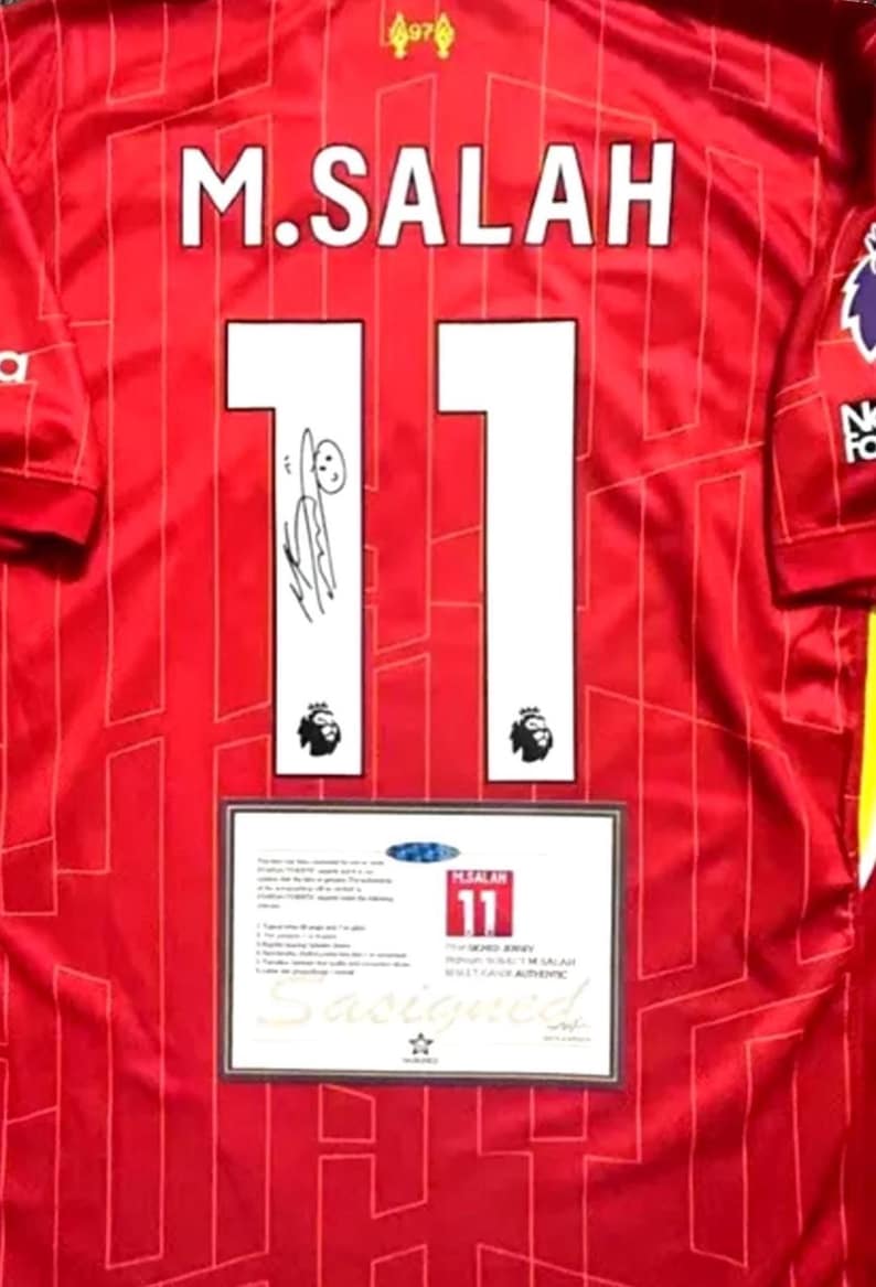 May include: A red soccer jersey with the name "M. SALAH" and the number "11" on the back. The jersey has a white signature on the number 1 and a white logo on the number 1. The jersey has a white tag on the bottom with the text "THE REAL DEAL" and "AUTHENTIC" in black.