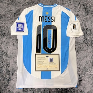 May include: A white and light blue soccer jersey with the number 10 and the name "MESSI" on the back. The jersey has the FIFA World Cup logo on the sleeve and a certificate of authenticity.