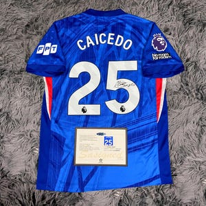 May include: A blue soccer jersey with the name "CAICEDO" and the number "25" printed in white. The jersey has a signature and the Premier League logo. A certificate of authenticity is displayed below the jersey.