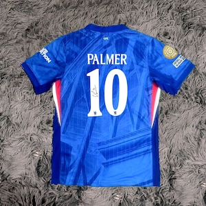 May include: A blue soccer jersey with the name "PALMER" and the number "10" in white. The jersey has a signature and team logos. The jersey is short-sleeved and has red and white accents on the sides.