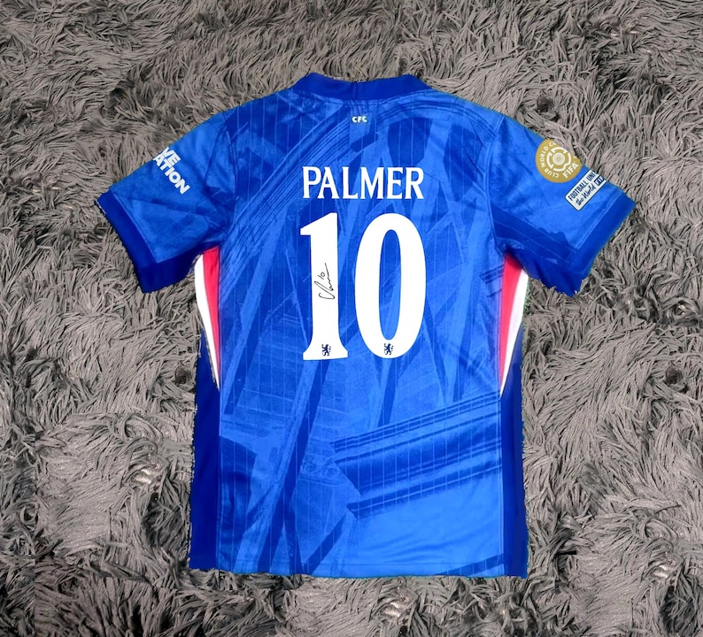 May include: A blue soccer jersey with the name "PALMER" and the number "10" in white. The jersey has a signature and team logos. The jersey is short-sleeved and has red and white accents on the sides.