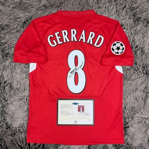 May include: Red soccer jersey with the name "GERRARD" and the number "8" in white. Features a soccer ball emblem on the sleeve and a certificate of authenticity. The jersey is signed.