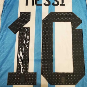 May include: Argentina soccer jersey with the name "MESSI" in black block letters above the number "10". The number is black with white polka dots. The jersey has blue and white stripes and a signature.
