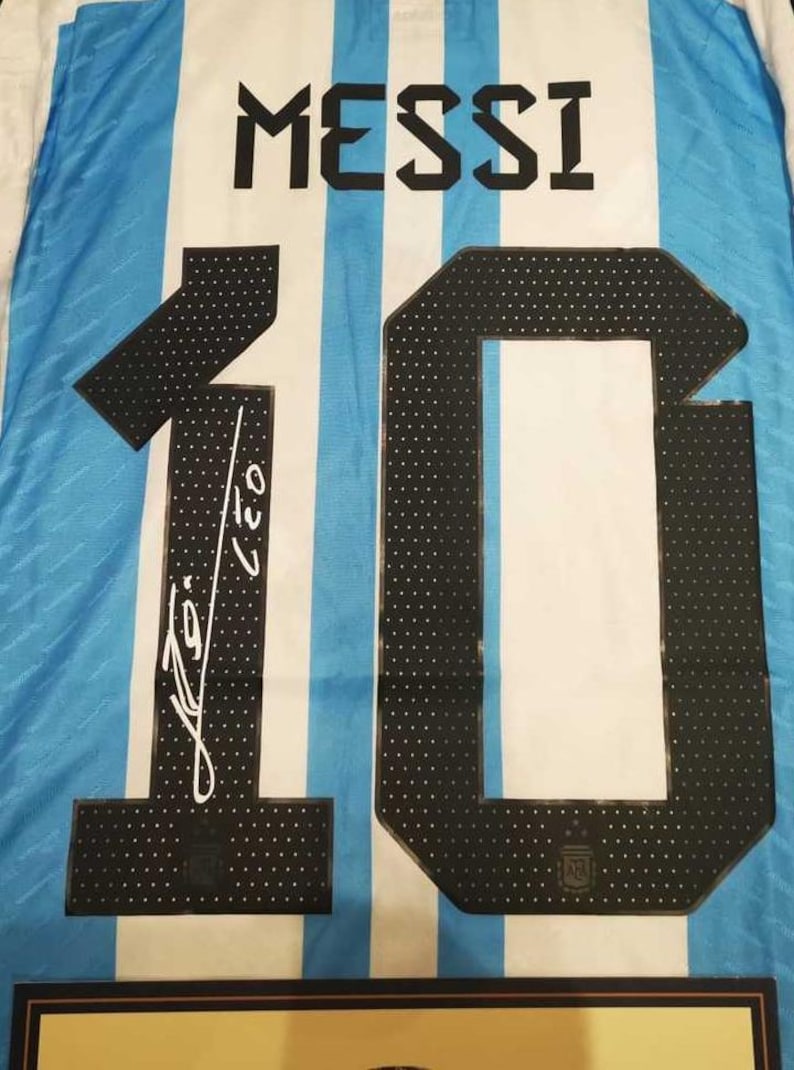 May include: Argentina soccer jersey with the name "MESSI" in black block letters above the number "10". The number is black with white polka dots. The jersey has blue and white stripes and a signature.
