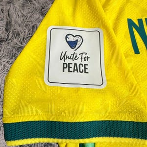 May include: A bright yellow athletic jersey with a textured geometric pattern, featuring a dark green hem and a white rectangular patch. The patch displays a blue heart icon above the black text Unite For PEACE. A partial green letter is visible.