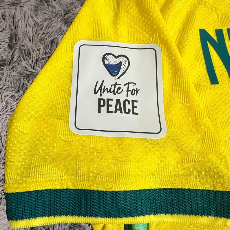 May include: A bright yellow athletic jersey with a textured geometric pattern, featuring a dark green hem and a white rectangular patch. The patch displays a blue heart icon above the black text Unite For PEACE. A partial green letter is visible.