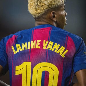 May include: A close-up of a blue and red soccer jersey with the name "LAMINE YAMAL" and the number "10" in yellow. The jersey has vertical red stripes and is worn by a person with blonde hair.