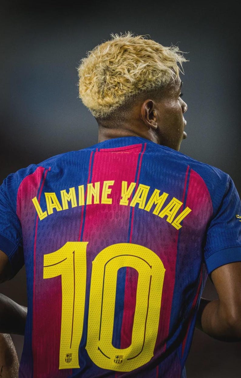 May include: A close-up of a blue and red soccer jersey with the name "LAMINE YAMAL" and the number "10" in yellow. The jersey has vertical red stripes and is worn by a person with blonde hair.