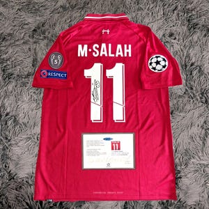 Może przedstawiać: Czerwona koszulka piłkarska Liverpool z nazwiskiem "M. SALAH" i numerem "11" na plecach. Koszulka ma biały kołnierz i białe akcenty. Posiada naszywkę "Respect" i logo Ligi Mistrzów na rękawach. Poniżej wyświetlany jest certyfikat.