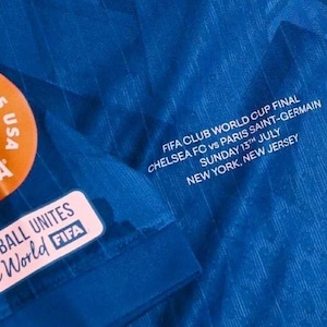 May include: Close-up of a blue soccer jersey with white text that reads "FIFA CLUB WORLD CUP FINAL CHELSEA FC vs PARIS SAINT-GERMAIN SUNDAY 13TH JULY NEW YORK, NEW JERSEY." The jersey also has FIFA patches.