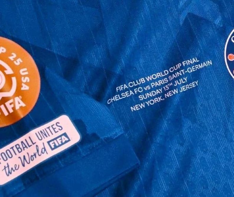 May include: Close-up of a blue soccer jersey with white text that reads "FIFA CLUB WORLD CUP FINAL CHELSEA FC vs PARIS SAINT-GERMAIN SUNDAY 13TH JULY NEW YORK, NEW JERSEY." The jersey also has FIFA patches.