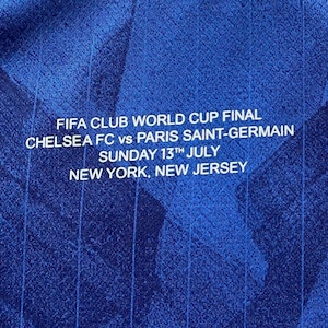 May include: Close-up of a blue jersey with white text. The text reads "FIFA CLUB WORLD CUP FINAL CHELSEA FC vs PARIS SAINT-GERMAIN SUNDAY 13TH JULY NEW YORK, NEW JERSEY". The jersey has a textured pattern.