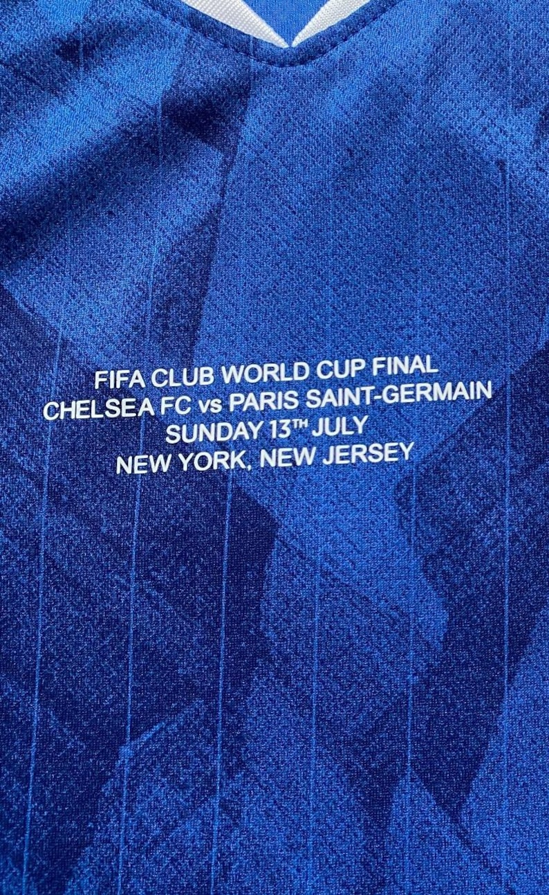 May include: Close-up of a blue jersey with white text. The text reads "FIFA CLUB WORLD CUP FINAL CHELSEA FC vs PARIS SAINT-GERMAIN SUNDAY 13TH JULY NEW YORK, NEW JERSEY". The jersey has a textured pattern.