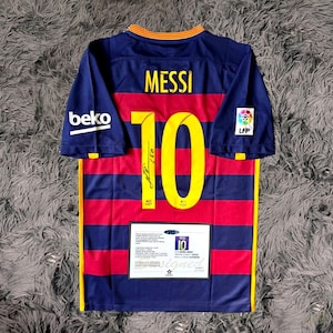 May include: A Barcelona soccer jersey with the name "MESSI" and the number "10" in yellow. The jersey is blue and red striped with a "beko" logo on the left sleeve and a "LFP" logo on the right sleeve. A signed certificate is displayed below the number.