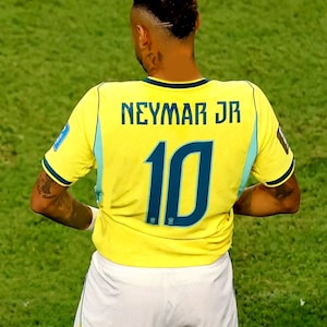 May include: A yellow soccer jersey with the name NEYMAR JR and the number 10 printed in dark blue on the back. The jersey features light blue side panels and is paired with white shorts. The athlete has a short, textured haircut with a shaved line.