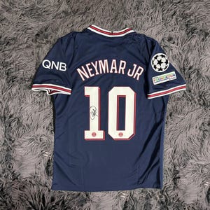 May include: Navy blue soccer jersey with the name "Neymar Jr" and the number "10" on the back. The jersey has white and red trim on the collar and sleeves, with the QNB logo on the left sleeve and a UEFA Foundation logo on the right sleeve.