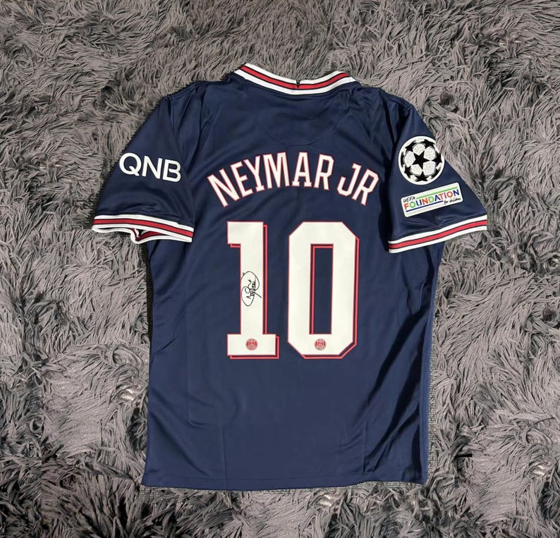 May include: Navy blue soccer jersey with the name "Neymar Jr" and the number "10" on the back. The jersey has white and red trim on the collar and sleeves, with the QNB logo on the left sleeve and a UEFA Foundation logo on the right sleeve.