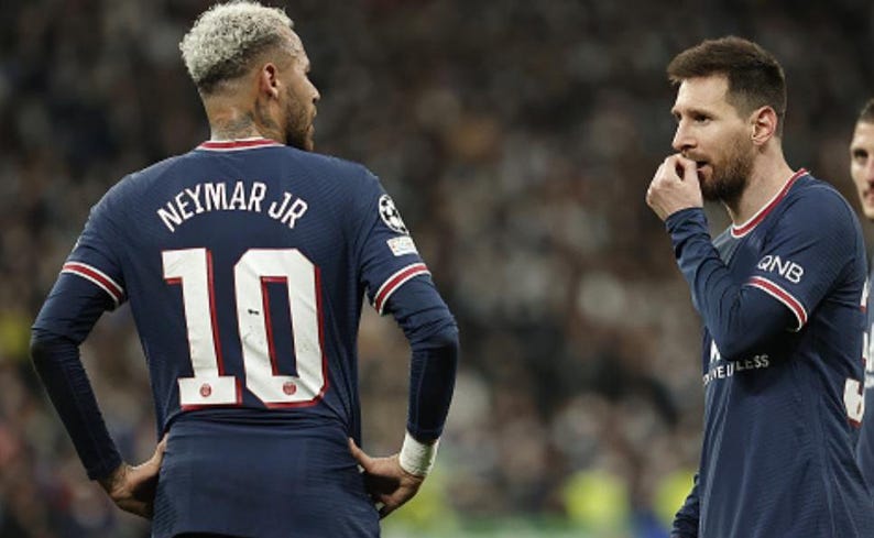 May include: Two soccer players in navy blue jerseys with red and white accents are seen on a field. One jersey displays "NEYMAR JR" and the number "10". The other player is biting his fingernails. The scene is set in a stadium.