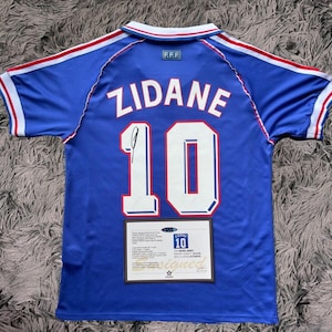 Pode incluir: Uma camisa de futebol azul com o nome "ZIDANE" e o número "10" em branco. A camisa tem listras vermelhas e brancas nas mangas e na gola. Uma pequena imagem emoldurada com o texto "ZIDANE 10 Signed" está na parte inferior.